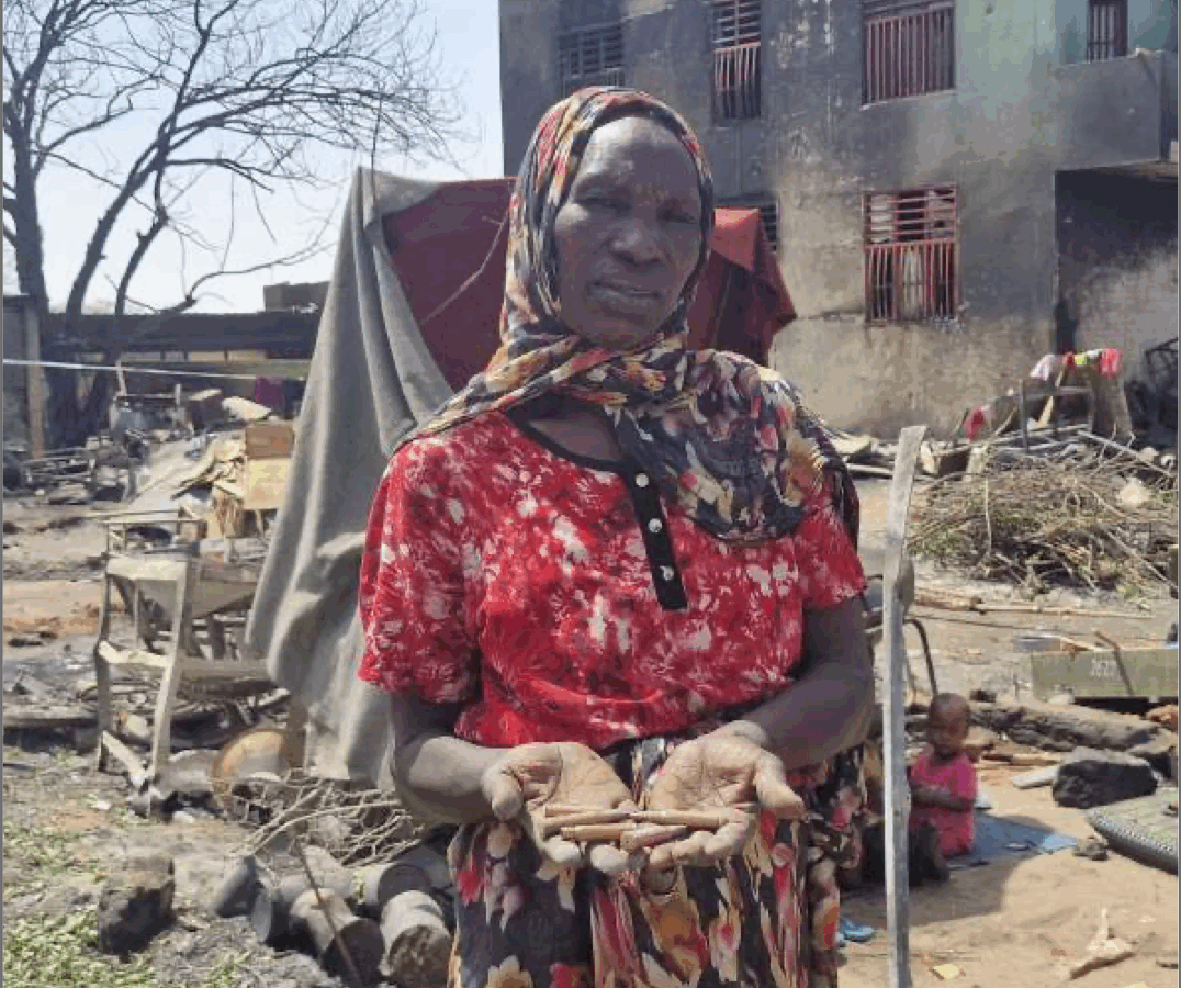 A Darfur woman standing in front of the ruins of a building, head covered in red dress.