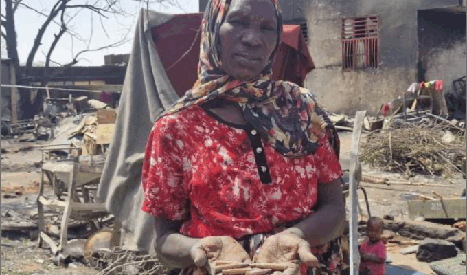 A Darfur woman standing in front of the ruins of a building, head covered in red dress.