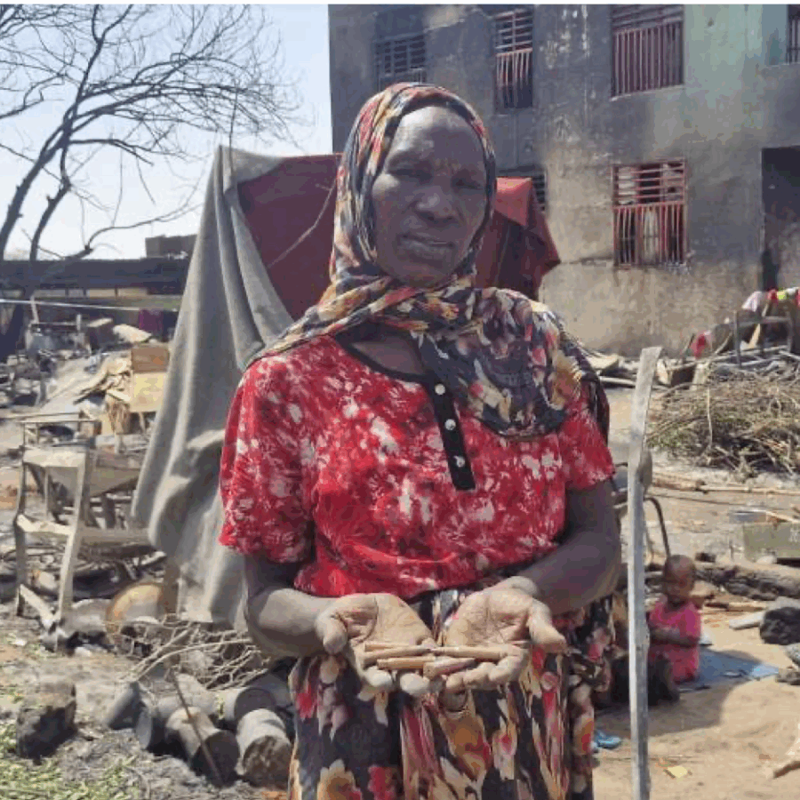 A Darfur woman standing in front of the ruins of a building, head covered in red dress.